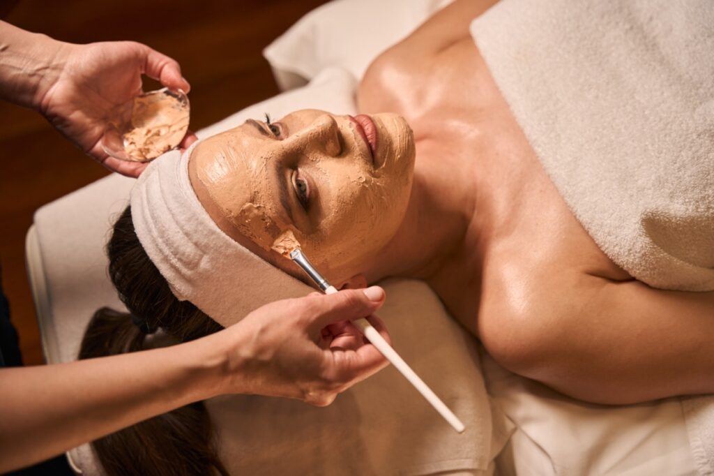 Kosmetikerin in einem Spa-Salon trägt mit einem Pinsel eine Maske auf das Gesicht einer ruhigen erwachsenen Frau auf.